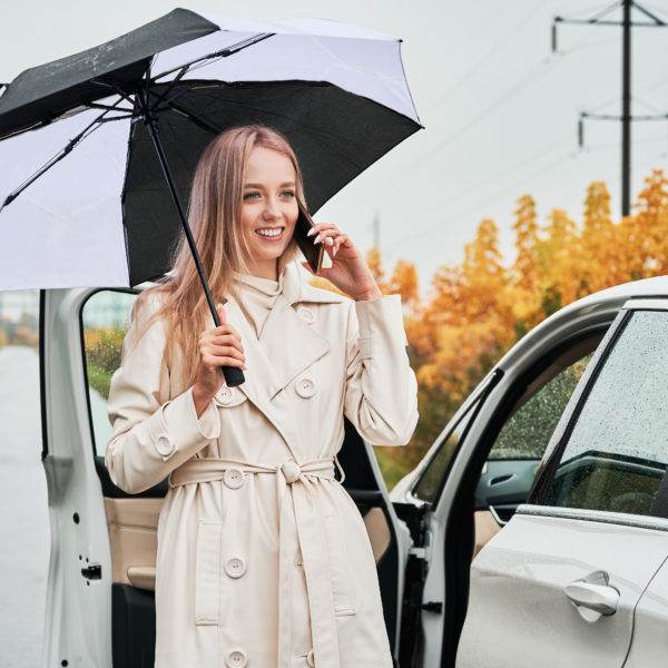 Portrait of smiling woman on a road near her stopped white car with punctured car tire. Female driver calling car service for help, holding umbrella.