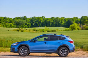 woman-blue-subaru-crosstrek-stranded-gravel-road-front-green-fields-distant-forest